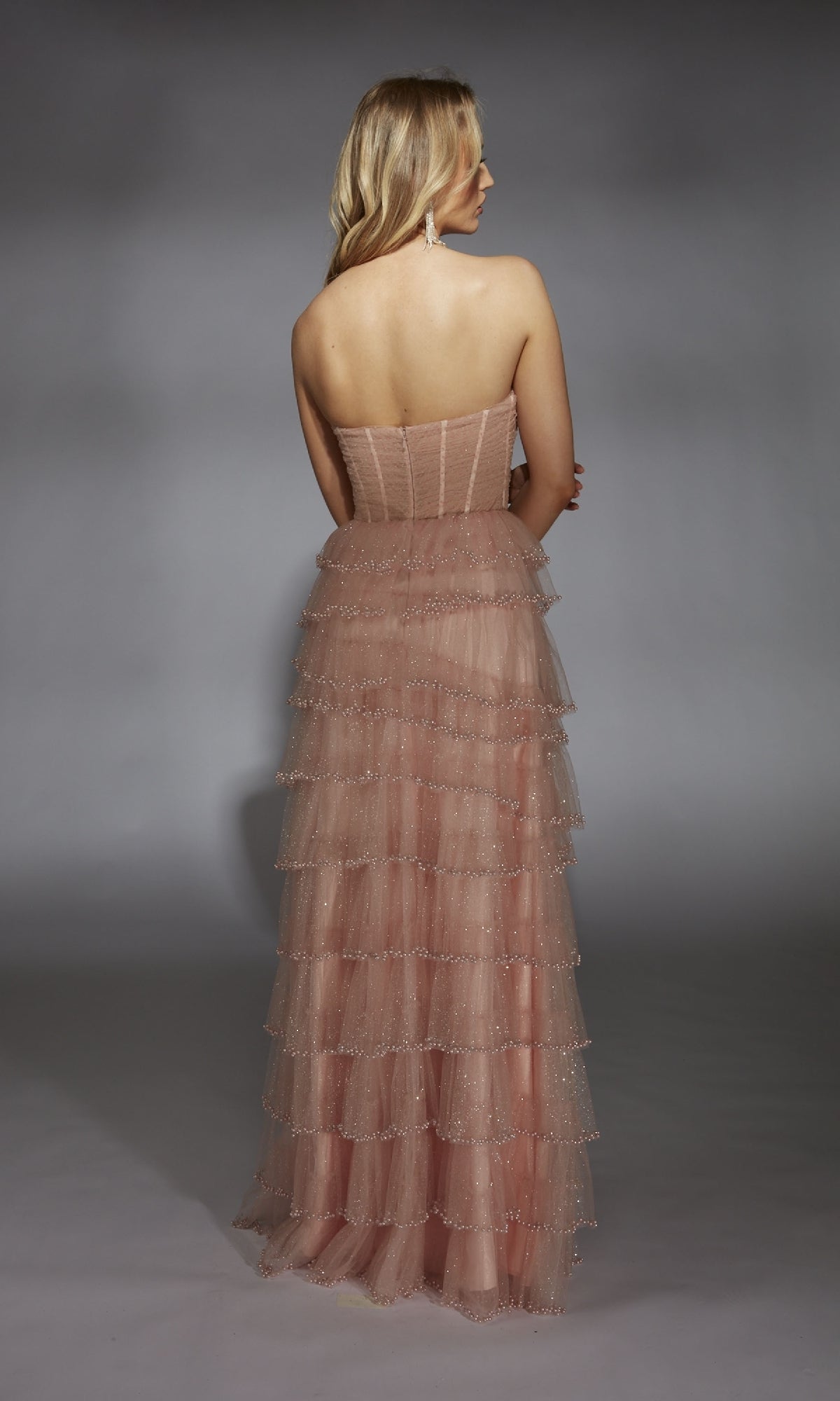 A woman faces away in the Alyce 61956 long prom dress, featuring a strapless glitter tulle design with a corset bodice and layered blush pink skirt, elegantly draped against a plain gray background.